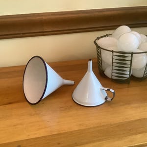 May include: Two white enamel funnels with dark rims. One funnel is lying on its side, while the other stands upright and features a small side handle. Beside them sits a dark wire basket filled with numerous smooth, round white spheres.