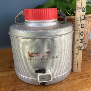 May include: A silver metal water cooler with a red lid and a white spigot. The cooler has a textured surface and the words "Cold  Drink Jug" are printed on the front.