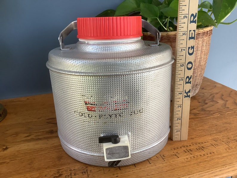 May include: A silver metal water cooler with a red lid and a white spigot. The cooler has a textured surface and the words "Cold  Drink Jug" are printed on the front.