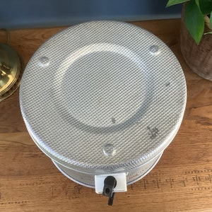 May include: A silver metal beverage dispenser with a textured surface and a black spigot. The dispenser is on a wooden surface.