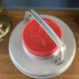 May include: A silver metal container with a red lid and a silver handle. The container has a textured surface.