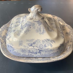 May include: A square ceramic butter dish with a lid featuring a blue and white floral transferware pattern. The lid has a sculpted handle at the top, and the base has a wide, flat rim with scalloped edges and matching floral details.