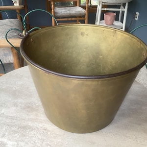 May include: A large, round, tapered brass bucket with a smooth, polished finish. The vessel features a dark, reinforced metal rim and two small, circular metal loops attached to the sides for handles. The interior shows a natural, aged patina.