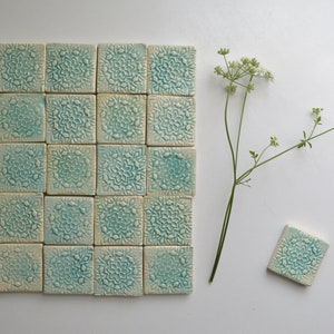 Puede incluir: Una disposición cuadrada de 25 baldosas de cerámica turquesa con un diseño floral. Una sola baldosa y una ramita de flores blancas están junto a las baldosas sobre una superficie blanca. Las baldosas miden unos 10 cm cuadrados.