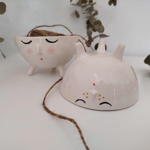Może przedstawiać: Dwie białe ceramiczne miski z namalowanymi twarzami. Jedna miska ma zamknięte oczy i różowy nos, druga ma zamknięte oczy i żółte kropki. Obie miski mają małe nóżki.