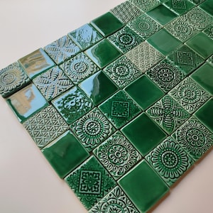 Puede incluir: Un conjunto de baldosas cerámicas cuadradas en tonos verdes y celeste. Cada baldosa presenta un diseño floral o geométrico en relieve. Las baldosas están dispuestas en diagonal, creando un mosaico visualmente atractivo.