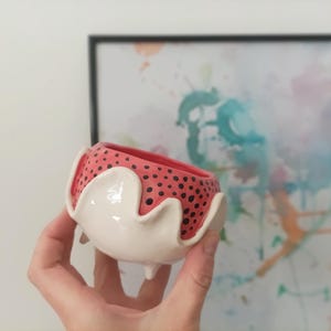 Può includere: Un vaso in ceramica bianca con un motivo a pois rosa e nero. Il vaso ha un bordo ondulato unico e tre piccoli piedini.