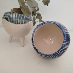 Może przedstawiać: Dwa ceramiczne doniczki z niebiesko-białymi paskami. Jedna doniczka ma trzy nóżki, a druga jest okrągła.