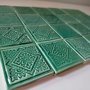Op de afbeelding: Een verzameling vierkante, smaragdgroene keramische tegels. Elke tegel heeft een verhoogd, sierlijk bloemen- en geometrisch patroon. De tegels zijn in een raster gerangschikt en tonen hun decoratieve potentieel.