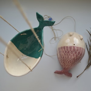 Op de afbeelding: Twee keramische hangende plantenbakken in de vorm van vissen. Eén is een lichtblauwe walvis met een witte buik en de andere is een roze vis met witte schubben. Beide hangen aan een touwtje.