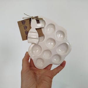Op de afbeelding: Witte keramische verfpalette met 10 individuele putjes, gevormd als een bloem. Een klein keramiek konijnenbeeldje is bevestigd aan de palette.