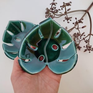 Może przedstawiać: Dwa ceramiczne naczynia w kształcie liści w odcieniach zieleni i turkusu. Naczynia mają błyszczące wykończenie z wyciętymi detalami i czerwonymi akcentami. Projekt przypomina roślinę Monstera. Dekoracyjna zastawa stołowa.
