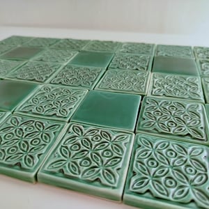 Op de afbeelding: Een verzameling vierkante, groene keramische tegels. Sommige tegels hebben een verhoogd bloemmotief, terwijl andere glad zijn. De tegels zijn in een rasterpatroon gerangschikt en creëren een decoratief oppervlak.