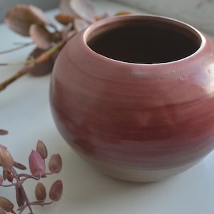 May include: A round, deep red ceramic planter with a smooth, glossy finish. The planter is empty and sitting on a white surface.