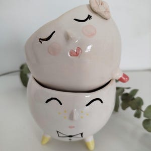Può includere: Due vasi in ceramica bianca a forma di viso. Il vaso superiore ha gli occhi chiusi, un naso rosa e una bocca a forma di cuore rosa. Il vaso inferiore ha gli occhi aperti, un naso rosa, un papillon e piedi gialli.