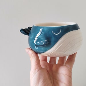 May include: A ceramic whale-shaped mug. The mug is primarily blue with white accents, featuring a fin-shaped handle and a smiling face design. The mug is held in a hand.