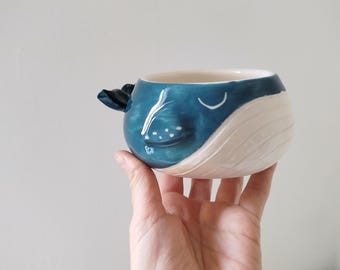 May include: A ceramic whale-shaped mug. The mug is primarily blue with white accents, featuring a fin-shaped handle and a smiling face design. The mug is held in a hand.