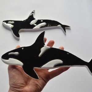 Op de afbeelding: Twee zwart-witte orka walvis decoraties. Eén wordt in de hand gehouden, de andere is erboven geplaatst. De orka's hebben witte markeringen en zijn gemaakt van een glad materiaal. De achtergrond is wit.