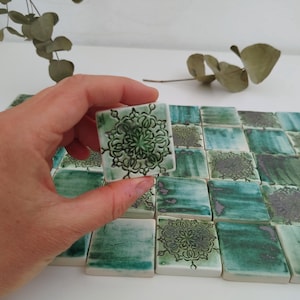 Op de afbeelding: Een verzameling groene en witte keramische tegels met een decoratief patroon. De tegels zijn in een rasterpatroon gerangschikt en hebben een licht getextureerd oppervlak.