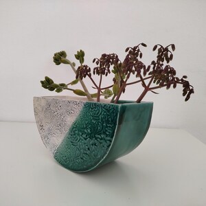 Op de afbeelding: Een halve cirkelvormige keramische pot met een witte en groene glazuur. De pot heeft een getextureerd oppervlak en is gevuld met een kleine groene plant met bruine stengels.
