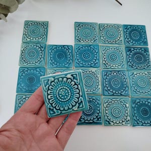 Peut inclure: Collection de carreaux de céramique turquoise carrés avec un motif floral. Les carreaux sont disposés sur une surface blanche, avec un carreau tenu dans une main. Le motif floral est une fleur centrale avec des pétales.