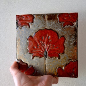 Könnte beinhalten: Quadratische dekorative Fliese mit einem roten Blumendesign auf silbernem und braunem Hintergrund. Die Fliese wird in einer Hand gehalten, wodurch die detaillierte Kunst hervorgehoben wird. Das Blumenmuster ist ein zentrales Element.