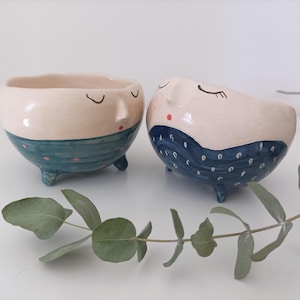 Puede incluir: Dos macetas de cerámica con forma de cara con los ojos cerrados y pequeñas patas. Las macetas están pintadas en un color beige claro con patrones de lunares azules y blancos.