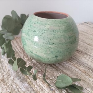 Op de afbeelding: Een groene keramische vaas met een gespikkeld glazuur. De vaas heeft een ronde vorm en een brede opening. Hij staat op een wit geweven kleed.