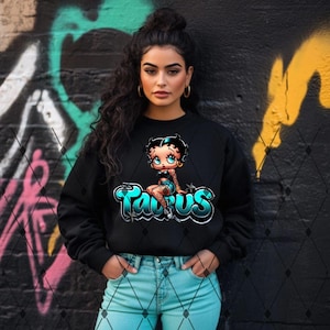 May include: Black crewneck sweatshirt featuring a cartoon illustration of a woman with the word "Taurus" in turquoise and silver. The design includes stars and a diamond pattern. The model is wearing light blue jeans.
