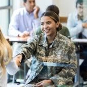 May include: A woman in a camouflage military uniform smiles and extends her hand in a handshake. She is seated in a classroom setting with other people at desks in the background.
