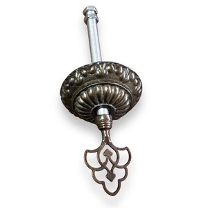 May include: A brass ceiling fixture with a decorative, ornate design. The fixture has a threaded rod and a key-shaped bottom.