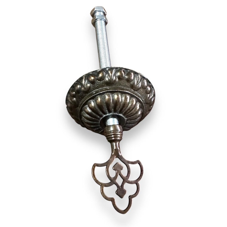 May include: A brass ceiling fixture with a decorative, ornate design. The fixture has a threaded rod and a key-shaped bottom.