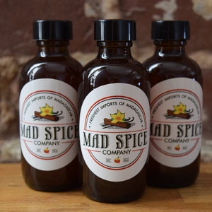 K&ouml;nnte beinhalten: Drei braune Glasflaschen mit schwarzen Deckeln und wei&szlig;en Etiketten. Die Etiketten zeigen einen roten und wei&szlig;en Kreis mit dem Text "Mad Spice Company" und einer Grafik eines Zimtst&uuml;cks und einer Vanilleschote. Der Text "Freshest Imports of Madagascar" ist auf dem Etikett aufgedruckt.