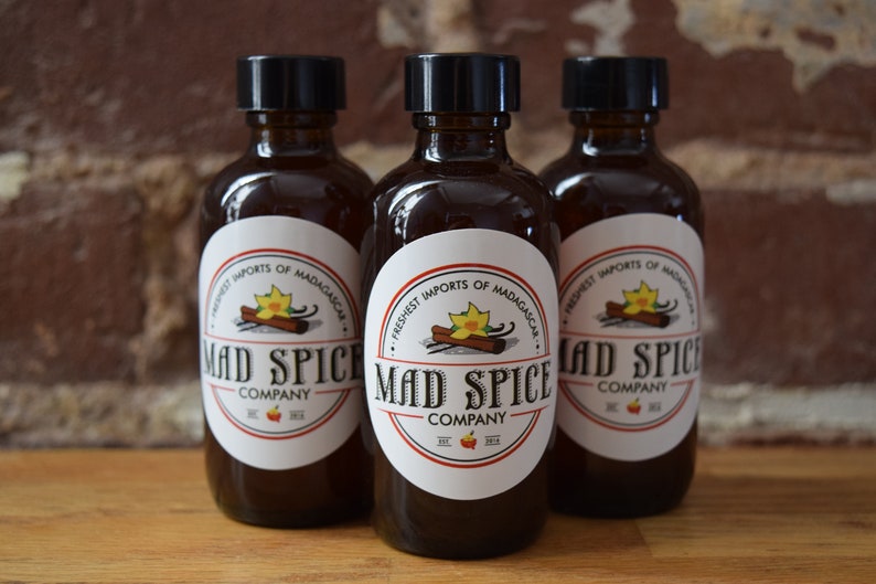 K&ouml;nnte beinhalten: Drei braune Glasflaschen mit schwarzen Deckeln und wei&szlig;en Etiketten. Die Etiketten zeigen einen roten und wei&szlig;en Kreis mit dem Text "Mad Spice Company" und einer Grafik eines Zimtst&uuml;cks und einer Vanilleschote. Der Text "Freshest Imports of Madagascar" ist auf dem Etikett aufgedruckt.