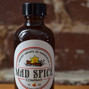 K&ouml;nnte beinhalten: Eine Glasflasche mit schwarzem Verschluss und einem wei&szlig;en Etikett. Das Etikett tr&auml;gt den Schriftzug "Mad Spice Company" und zeigt eine gelbe Blume, Zimtstangen und Vanilleschoten. Der Text auf dem Etikett lautet au&szlig;erdem "Freshest Imports of Madagascar" und "Est. 2014".