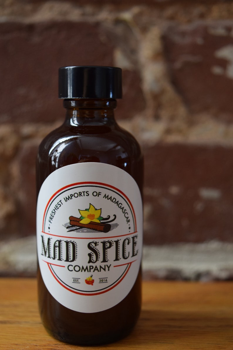 K&ouml;nnte beinhalten: Eine Glasflasche mit schwarzem Verschluss und einem wei&szlig;en Etikett. Das Etikett tr&auml;gt den Schriftzug "Mad Spice Company" und zeigt eine gelbe Blume, Zimtstangen und Vanilleschoten. Der Text auf dem Etikett lautet au&szlig;erdem "Freshest Imports of Madagascar" und "Est. 2014".