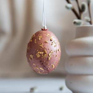 May include: A pink Easter egg with gold leaf accents hanging from a white string. The egg is decorated with a scattered pattern of gold leaf flakes.