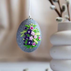 May include: A grey Easter egg with a floral design. The egg is decorated with pink and purple flowers and green leaves. The egg is hanging from a white string.