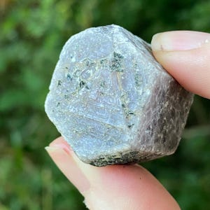 May include: A rough, uncut gemstone with a multifaceted, geometric shape. The stone has a pale purple hue with darker inclusions and a textured surface. The bottom edge is a darker, almost black color.