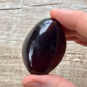 May include: A smooth, polished, dark purple gemstone in the shape of an oval. The stone is being held in a hand.