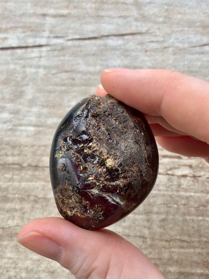 May include: A dark red, brown, and gold amber stone with a rough, natural texture. The stone is oval-shaped and has a smooth, polished surface.