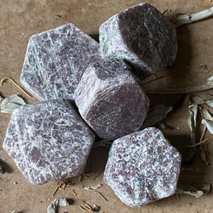 May include: A collection of four raw, hexagonal ruby crystals with a deep maroon and dusty purple hue. Each stone features a rough, textured surface with visible crystalline structures and natural matte finishes, arranged in a cluster formation.