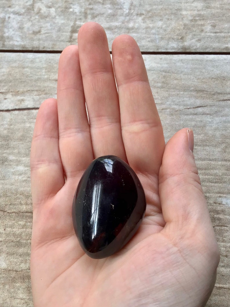 May include: A smooth, dark brown, polished gemstone in the shape of an oval. The stone is being held in a person's hand.