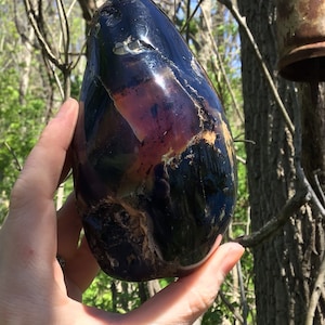 May include: A large, smooth, polished, black and purple agate stone with a unique, swirling pattern. The stone is held in a hand.