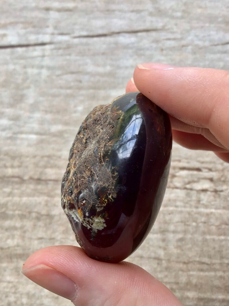 May include: A dark red, oval-shaped piece of amber with a rough, textured surface. The amber has a glossy finish and appears to have some inclusions.
