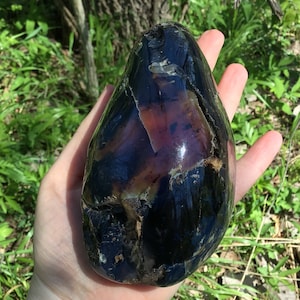 May include: A large, smooth, polished, dark purple and black gemstone with iridescent streaks. The gemstone is shaped like an egg and has a glossy finish.