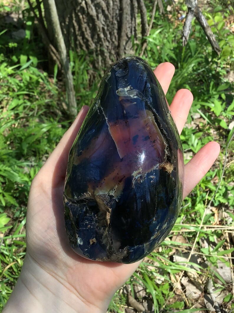 May include: A large, smooth, polished, dark purple and black gemstone with iridescent streaks. The gemstone is shaped like an egg and has a glossy finish.