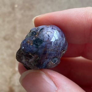 May include: A natural, raw, blue-purple gemstone with dark inclusions. The stone has a rounded shape and a slightly rough texture, held between fingers. The gem's surface reflects light, showcasing its color variations.