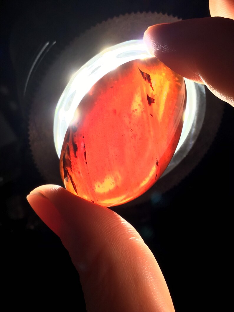May include: A smooth, oval-shaped, red gemstone with dark brown streaks and swirls. The gemstone is being held between two fingers.