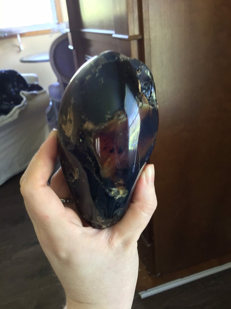 May include: A large, dark amber stone with a unique, swirling pattern of brown, black, and purple hues. The stone is smooth and polished, with a glossy finish.